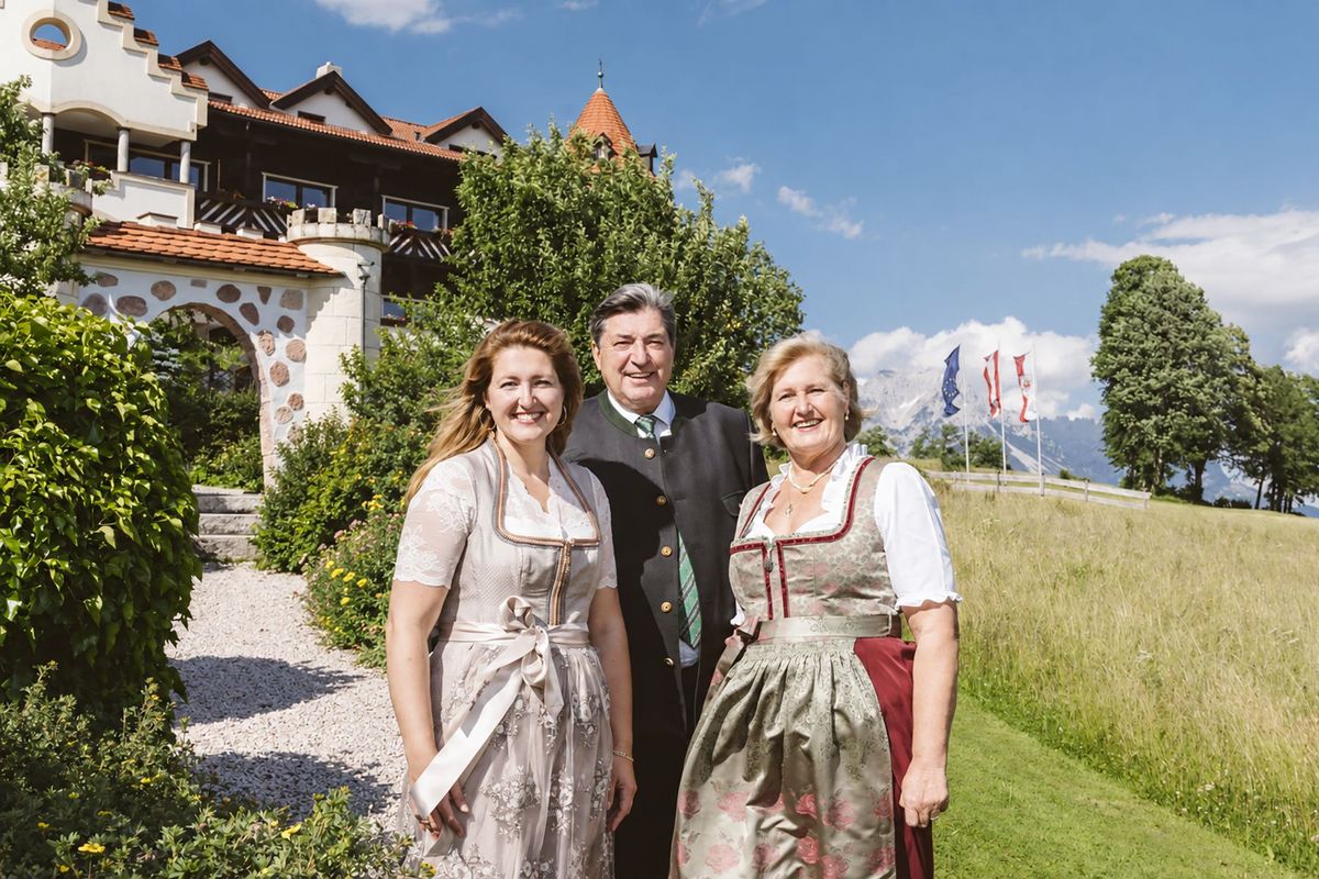 Carina, Karin und Hans – Gastgeber des Hotels AlpenSchlössl in Söll Carina, Karin und Hans, die Gastgeber des Hotels AlpenSchlössl in Söll, stehen in traditioneller Tracht vor der alpinen Landschaft.