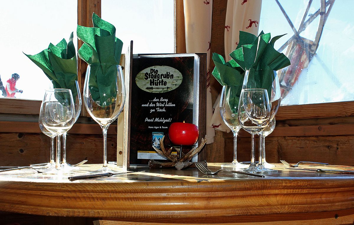 Gedeckter Tisch auf der Stoagrub’n Skihütte Gedeckter Tisch mit Weingläsern, grünen Stoffservietten und Speisekarte am Fenster der Stoagrub’n Skihütte.